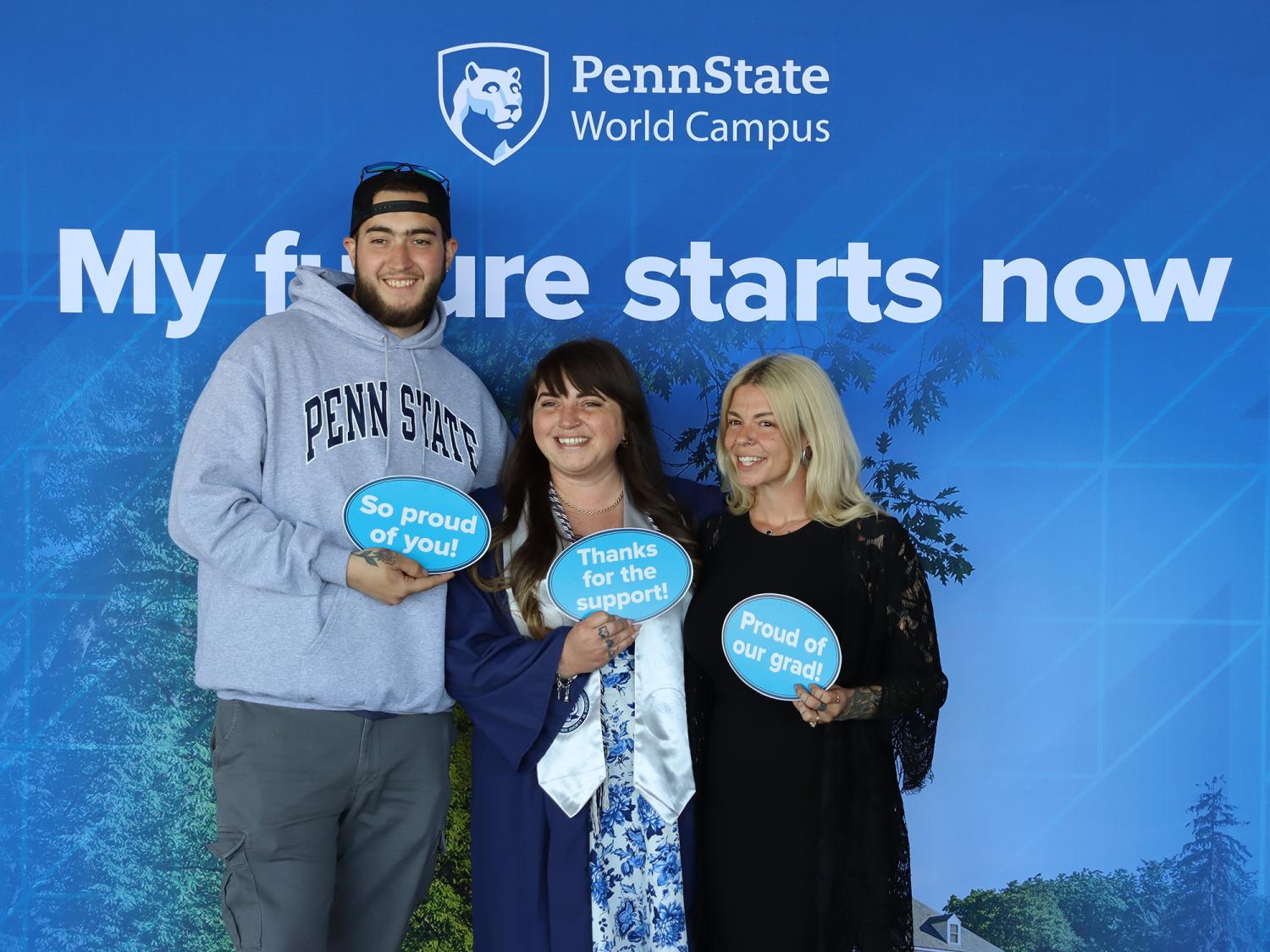 Three people holding signs standing in front of a Penn State photo background.