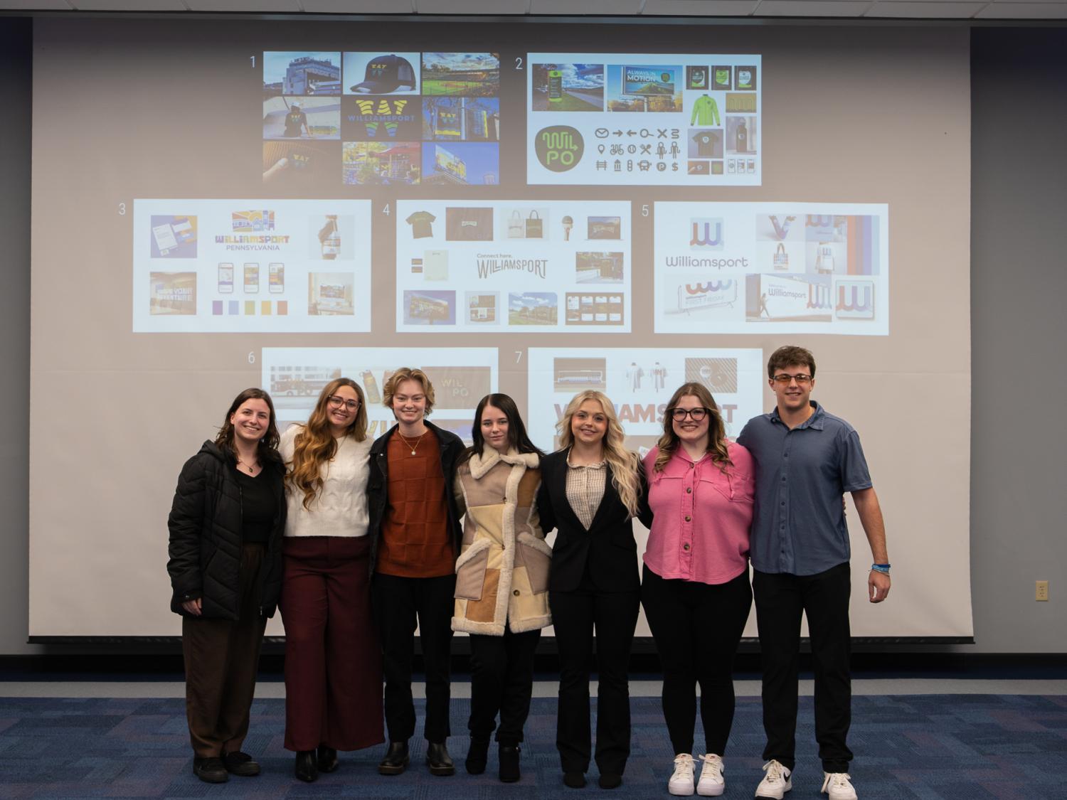 Seven students stand in front of a projection screen for a photo