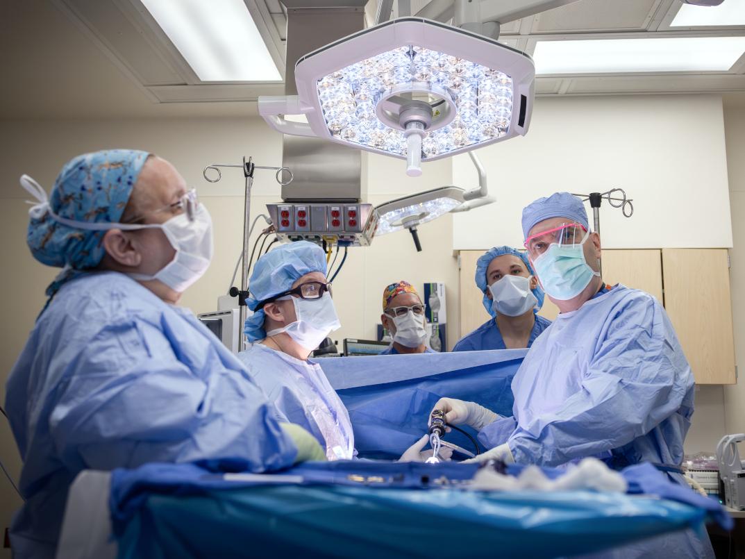 A surgical team wearing blue gowns, masks, and caps performs a procedure in a brightly lit operating room, standing around a patient draped in sterile blue sheets beneath an overhead surgical light.