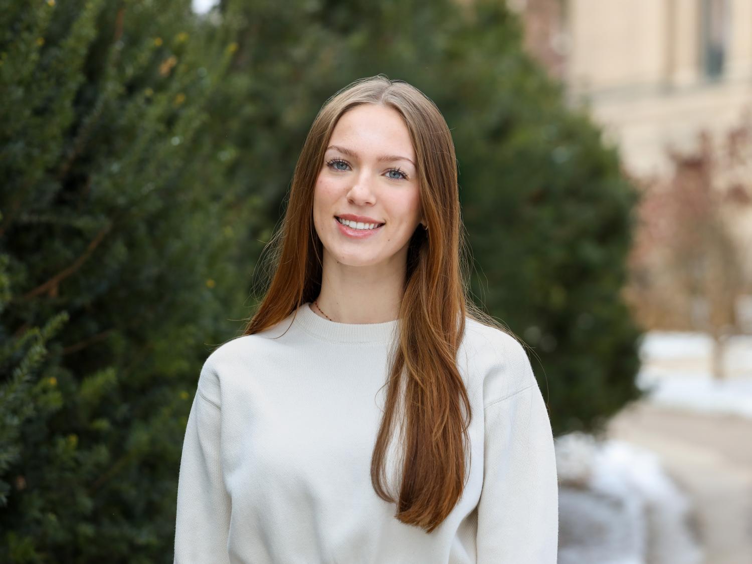 Alexis Wilson stands in a white sweater in front of green evergreen trees outside Sparks Building.
