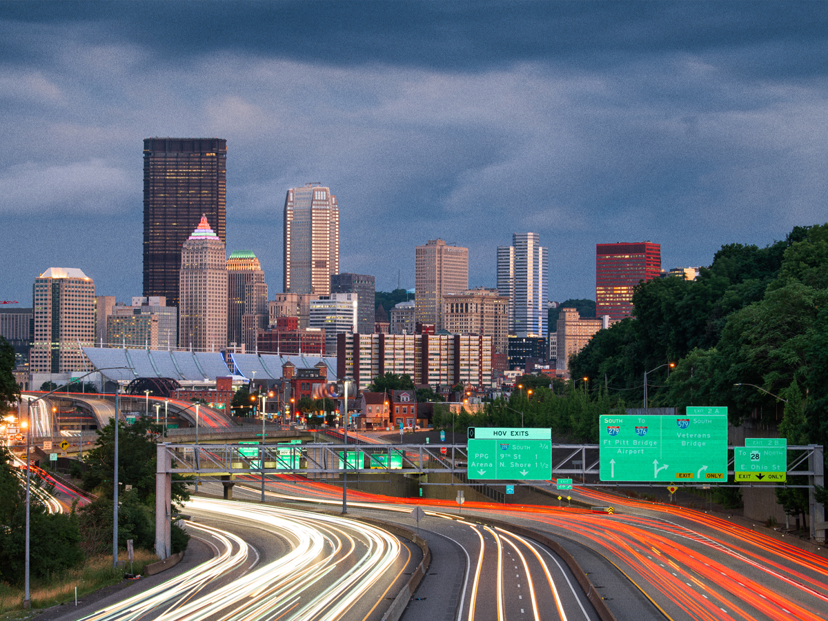 A photograph of Pittsburgh's skyline; in the foreground, cars drive on the highway leading into the city.