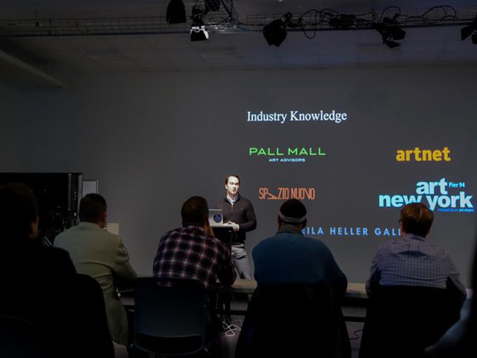 A man with dark hair and a black shirt presents in front of a projector screen to a panel of judges