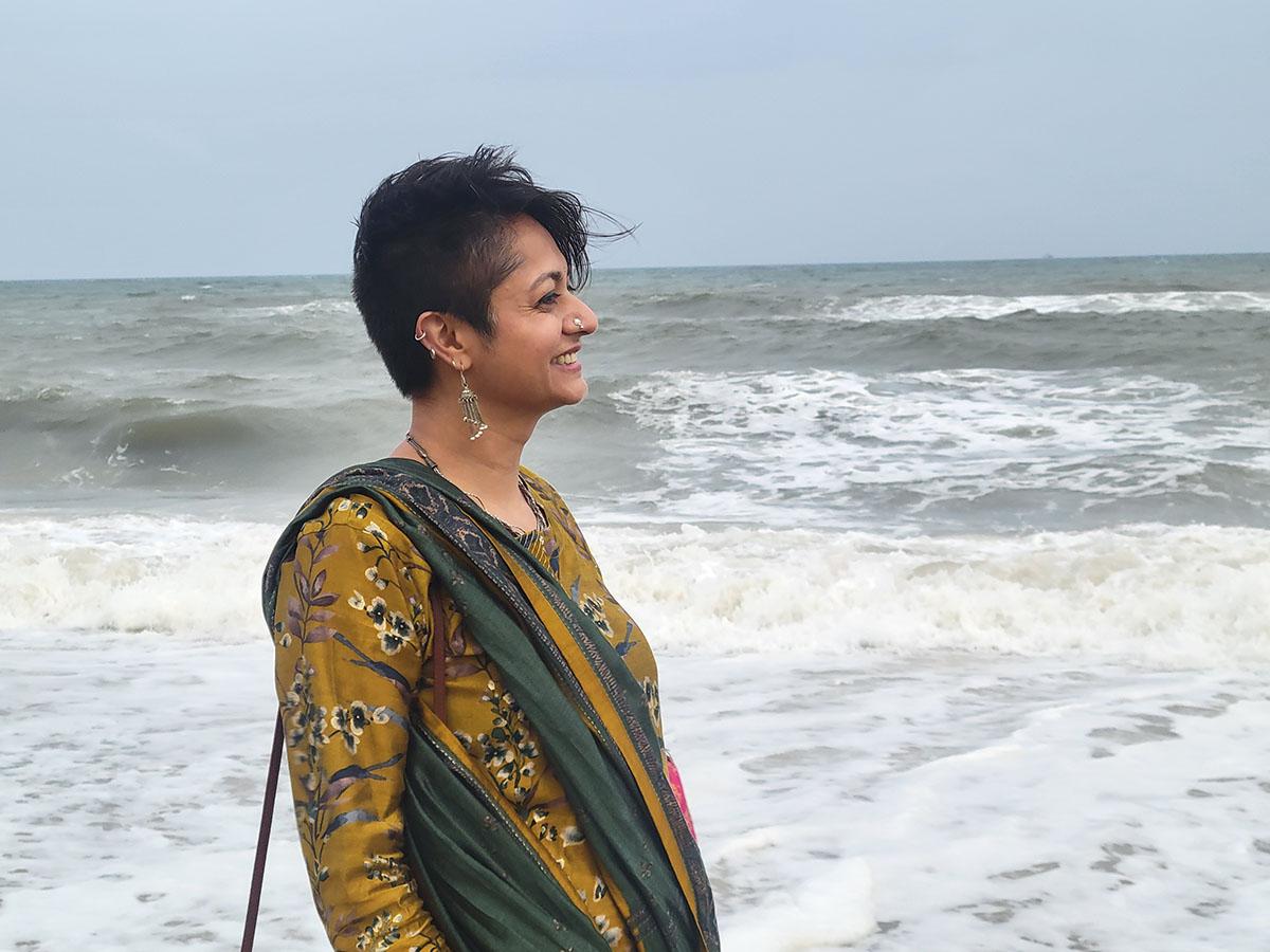 Dr. Vasudevan with short hair stands by the ocean, wearing a yellow floral top and green shawl.