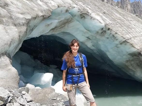 Elisabeth Clyne, a person with a long brown ponytail stands next to a glacier wearing a blue t-shirt, khaki shorts and a hiking backpack.