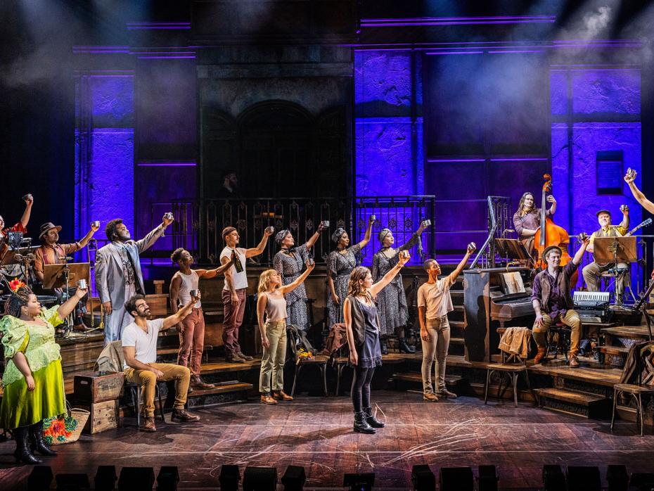 A diverse cast of actors toasts a man standing on a stool.