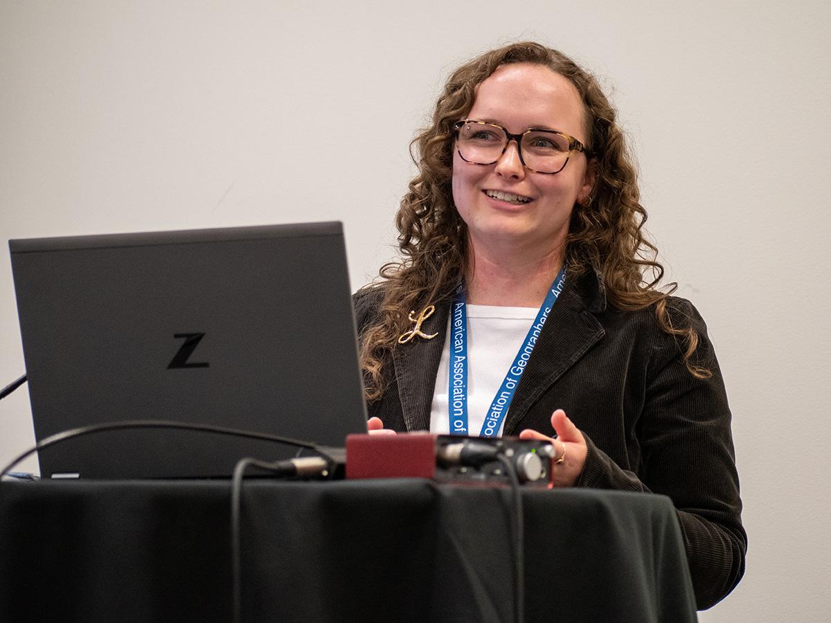 Lucy Thompson who has curly hair and glasses smiles while giving a presentation. They are standing behind a podium with a laptop. 