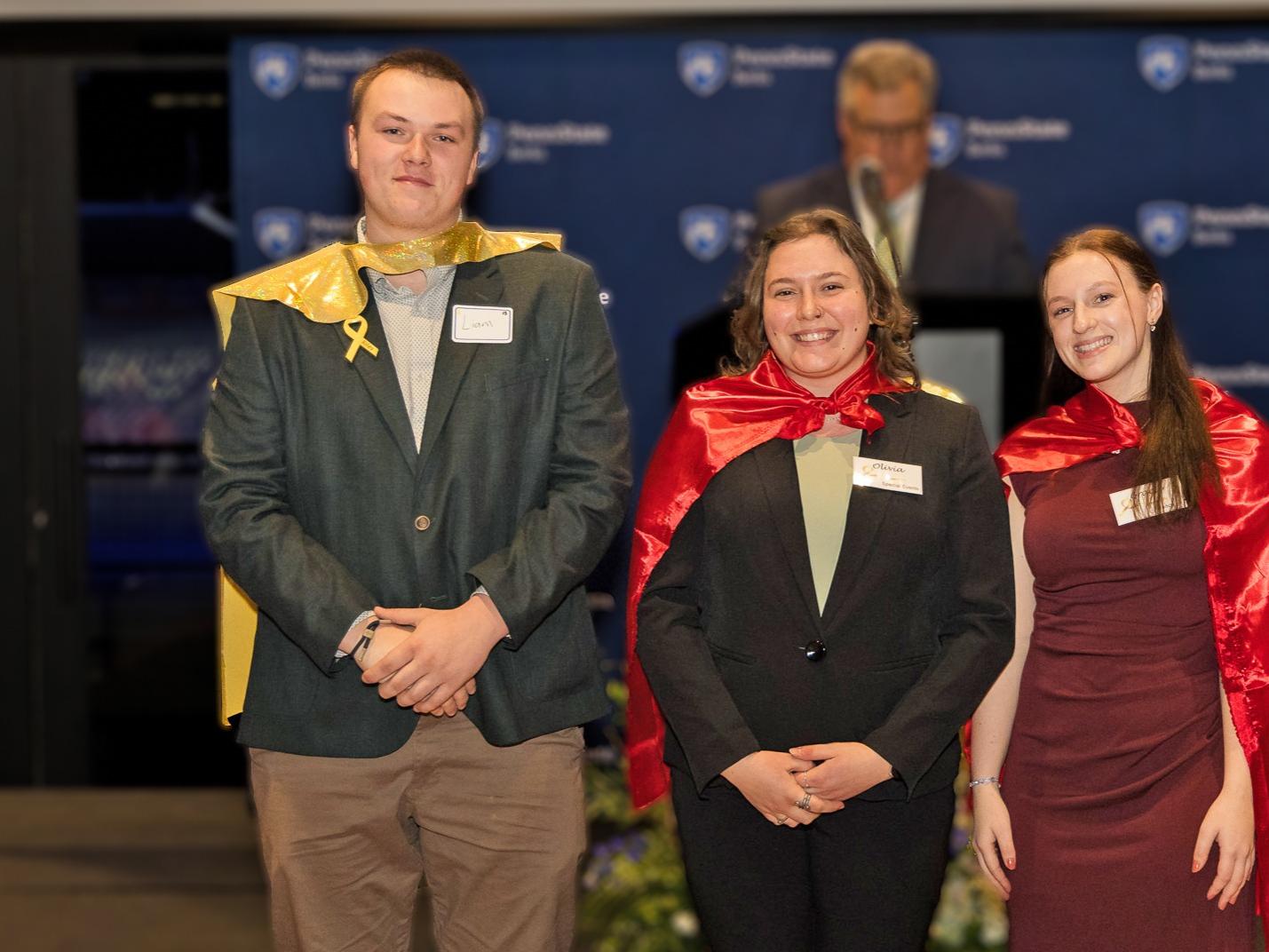 three students stand in a line wearing capes pose for a photo