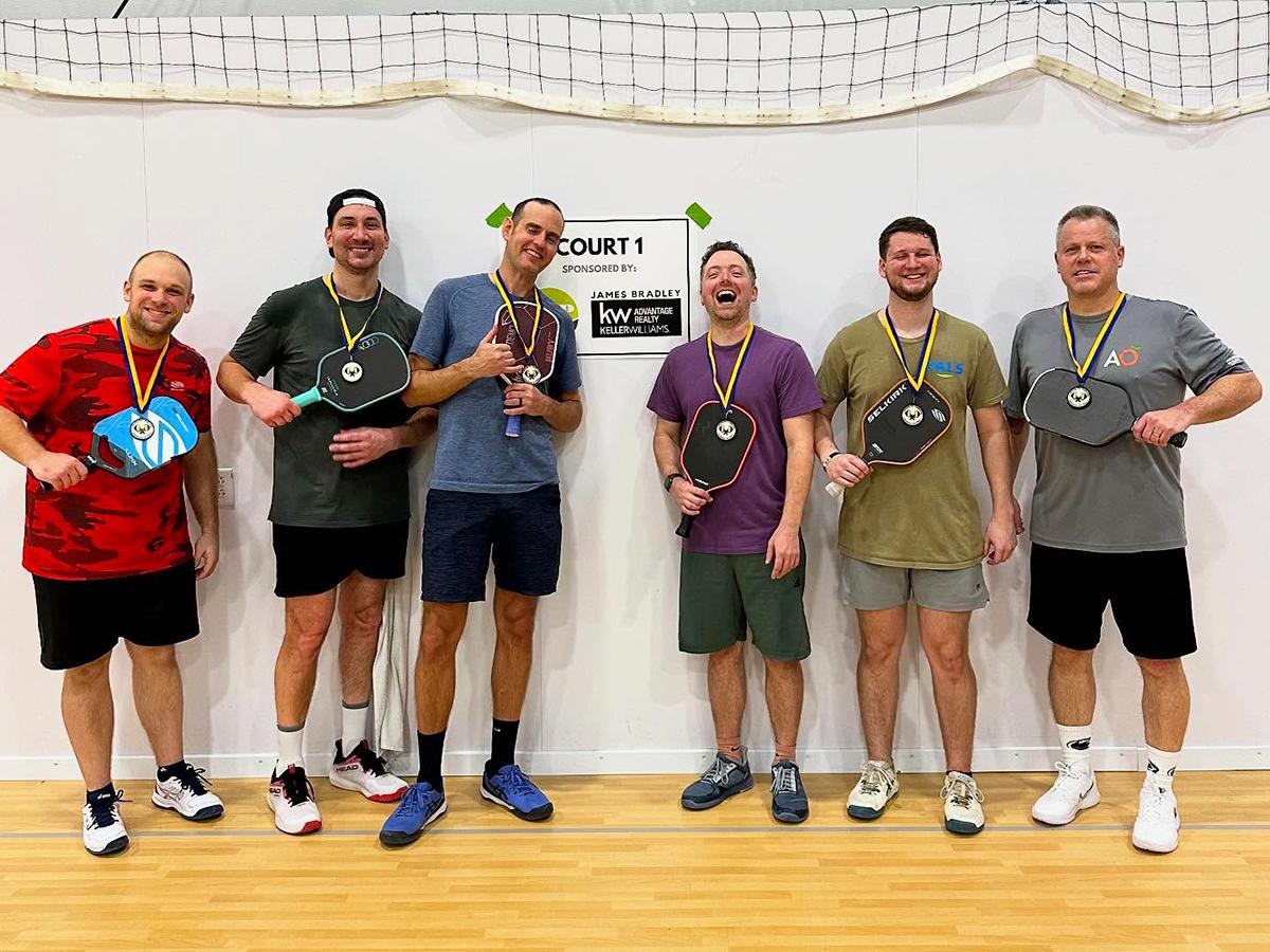 A group of men standing indoors on a gym floor, smiling and posing with pickleball paddles while wearing medals, suggesting a friendly competition or tournament.