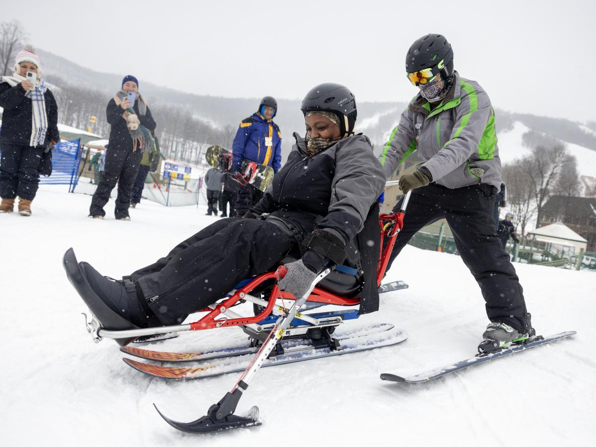 A woman is seated on an adaptive sled, while a ski instructor in winter clothing and on skis holds onto the back of the sled. They are on snow, and a few people look on from the background.