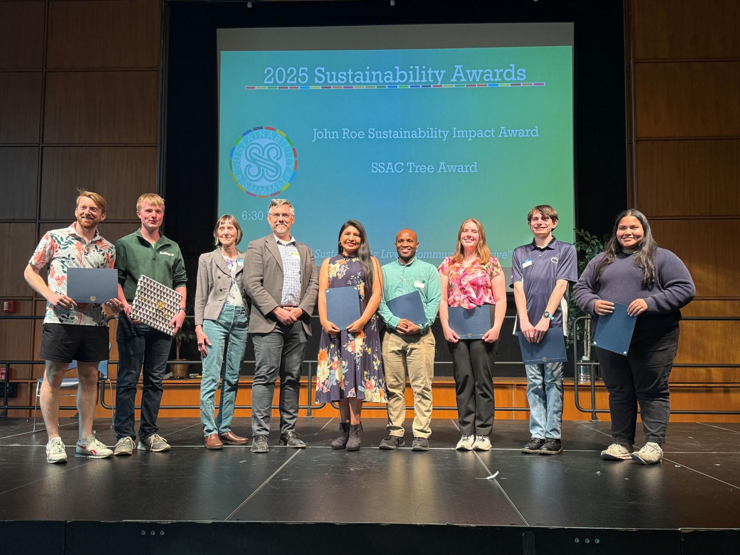 A group of nine students and award presenters on a stage displaying the John Roe Sustainability Impact Awards.