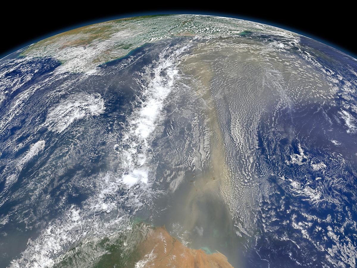 Satellite view of Earth showing the Sahara Desert in the foreground, with a vast plume of dust extending over the Atlantic Ocean under a cloudy sky.