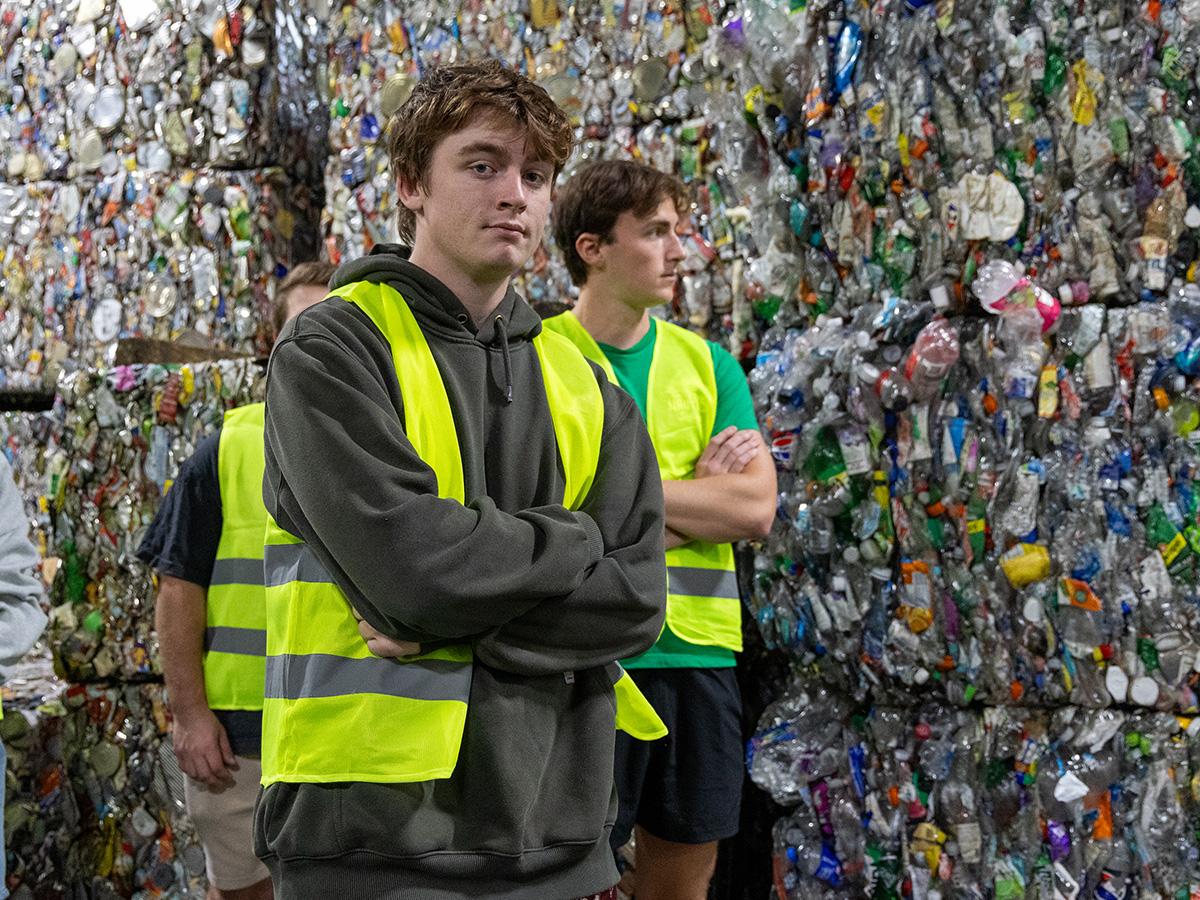 Students in reflective vests stand in front of large piles of compacted plastic bottles at a recycling facility.