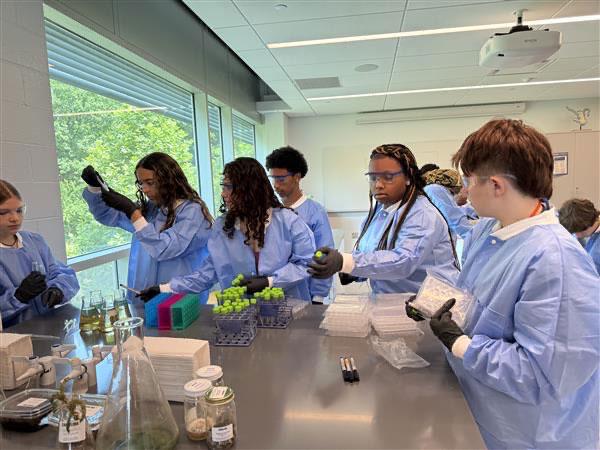 Students in protective lab gear gather around a table of vials and flasks