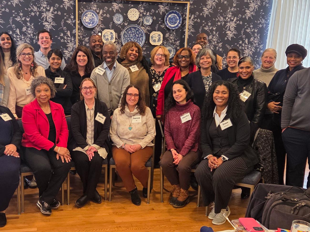 A large group of symposium attendees pose together for a group photo in a room with patterned wallpaper and a decorative display of plates on the wall