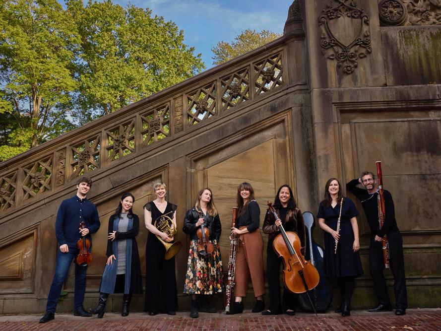 A group of eight chamber musicians stand against a stone wall.