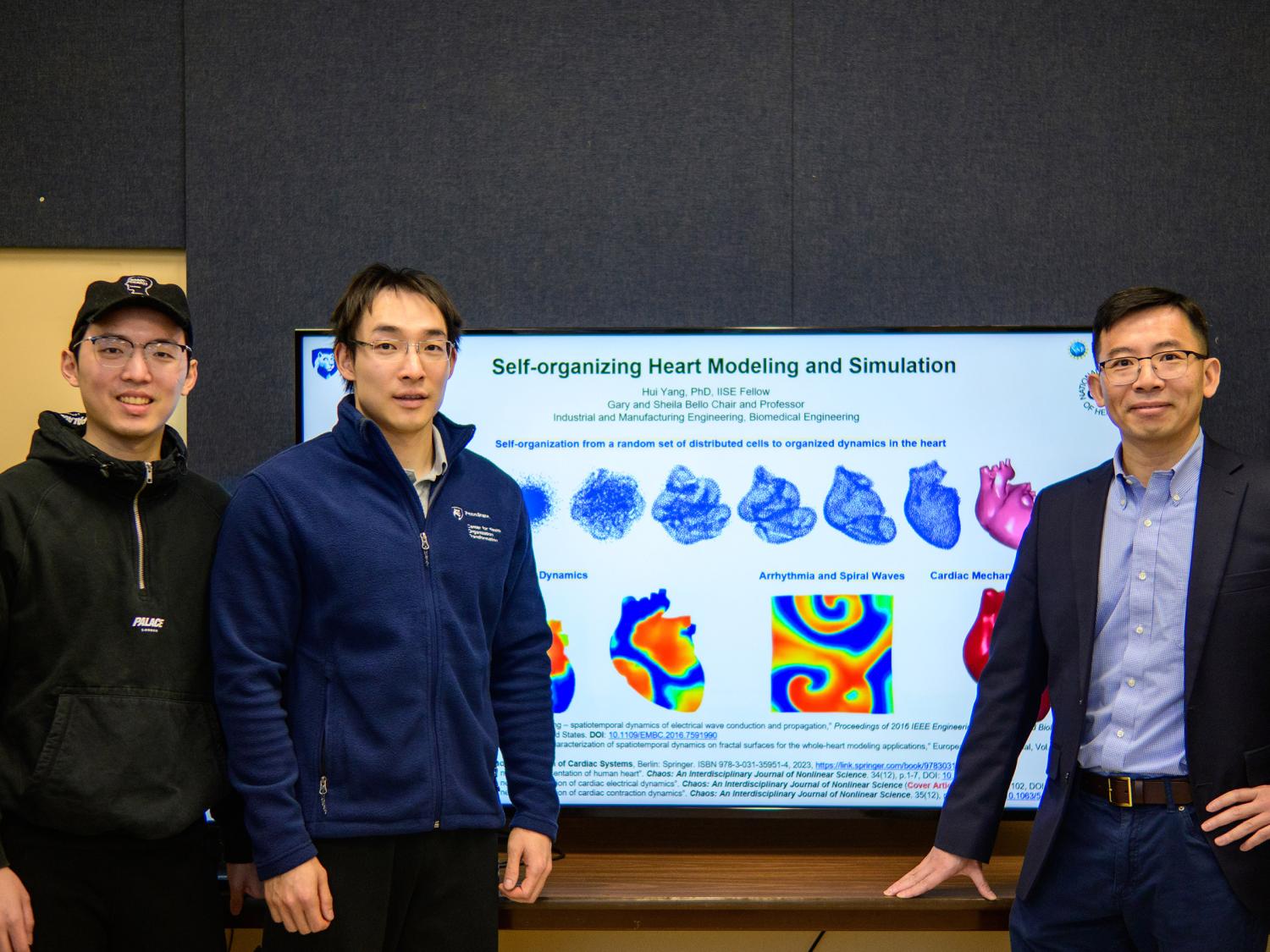 Three men pose for a photo in front of a television that reads "Self-organizing Heart Modeling and Simulation" with a series of graphics of graphics showing the heart in different phases of self-organization, with electrical maps of a heart beneath it. 