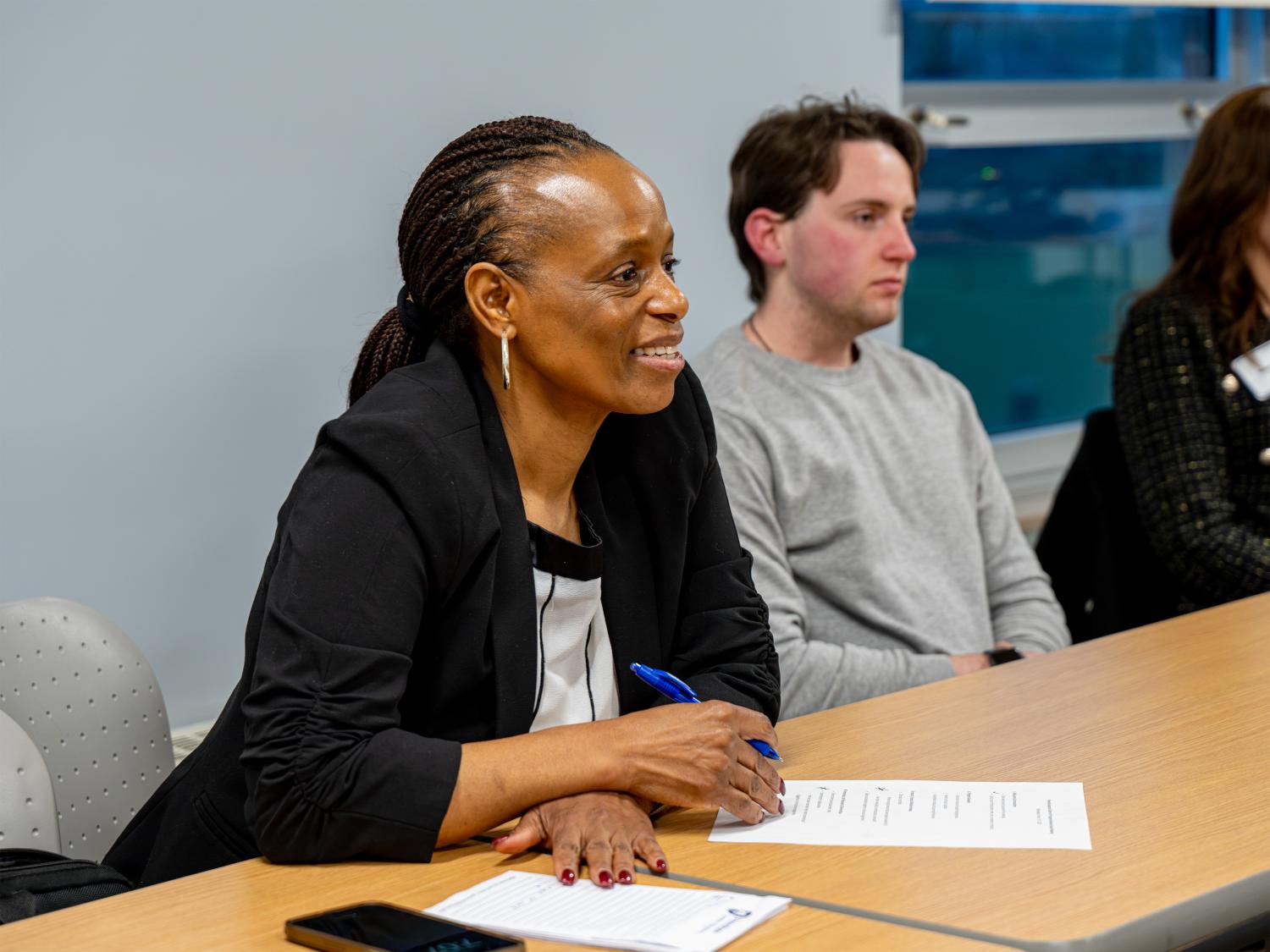 Two people sit at a conference table with papers and a pen during a meeting, while additional participants are seated nearby in a classroom or conference room setting with windows along the wall.