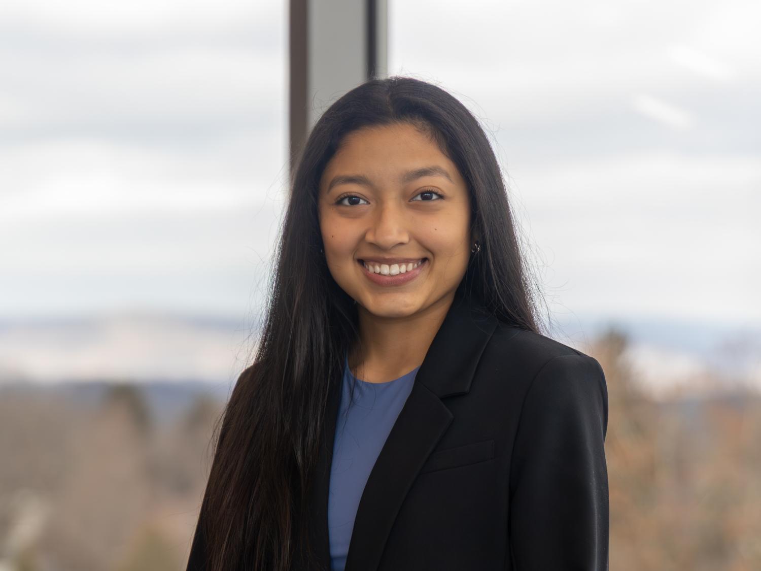 Ashley Carmona wears a black blazer over a blue shirt while standing in front of a window inside Welch Building.