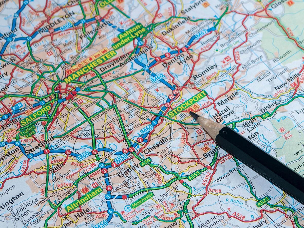 A detailed road map showing Manchester, Stockport, and surrounding areas, with roads highlighted in red and green. A pencil points to Stockport.
