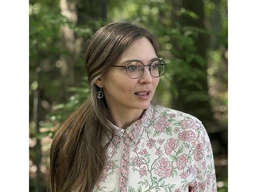 Danielle Rye wearing a white blouse with flowers while standing in front of trees