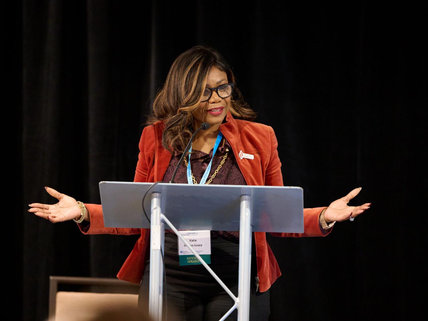 Woman in a red blazer speaking at a podium with a black backdrop during a conference.