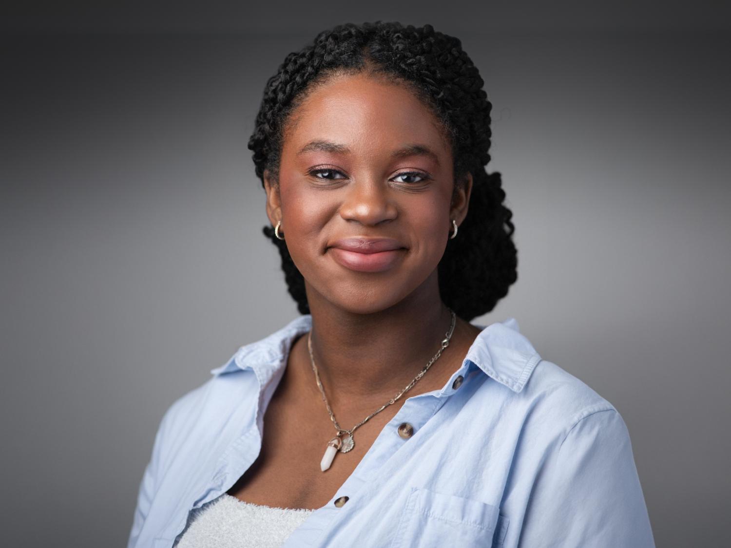 Christina Ellis poses for a headshot while wearing a light blue button-up shirt and a crystal pendant necklace against a neutral background.