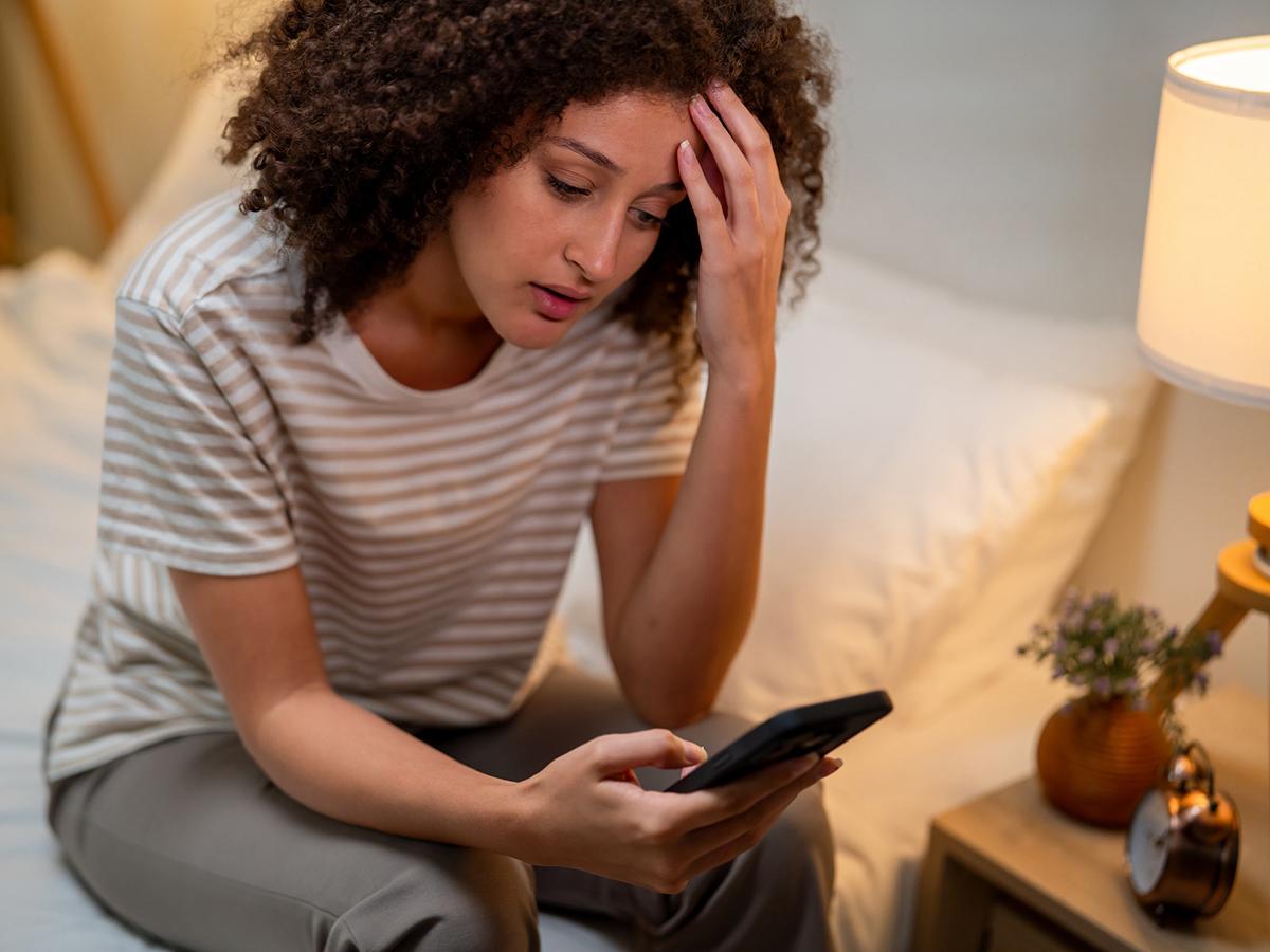 Stressed woman uses mobile phone as she sits on bed at night. 