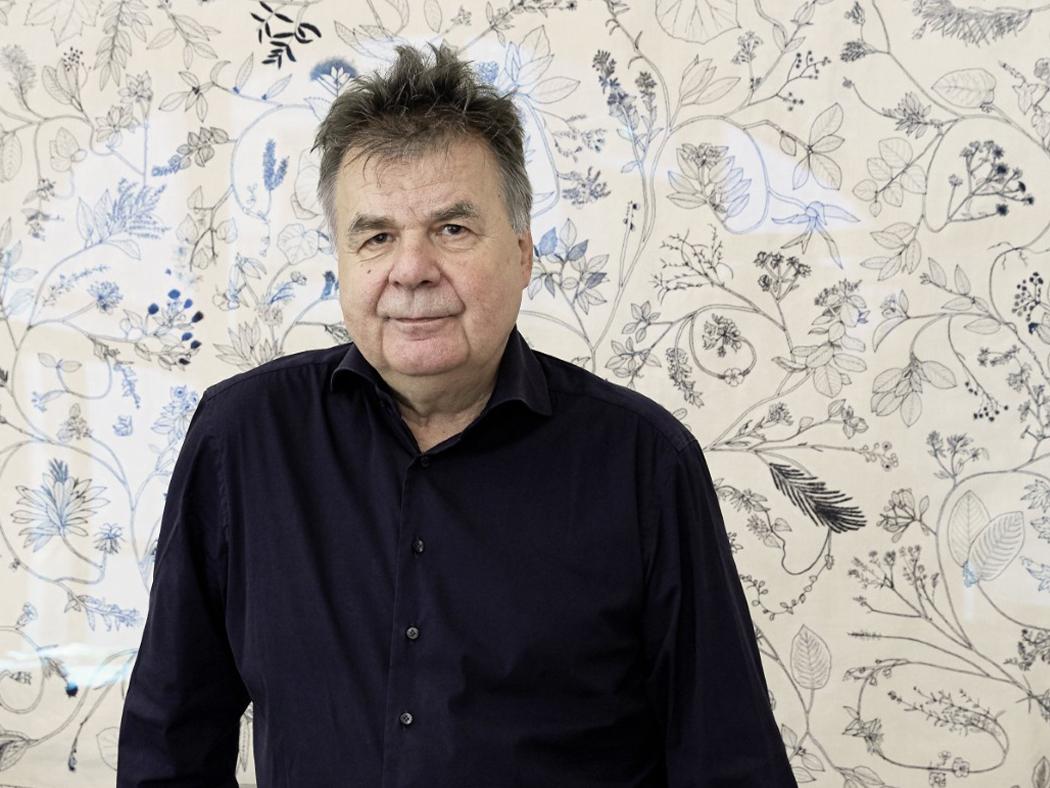 Man with grayish spiky hair and black button-down shirt standing in front of wall with white and black floral wallpaper