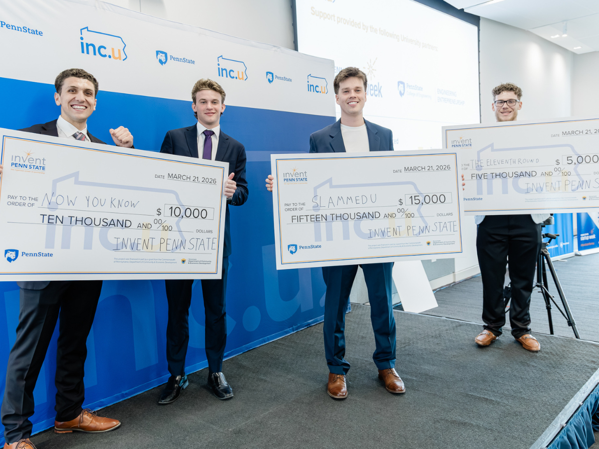 Four students stand on a stage in front of Inc.U signage, students hold up three different oversized checks