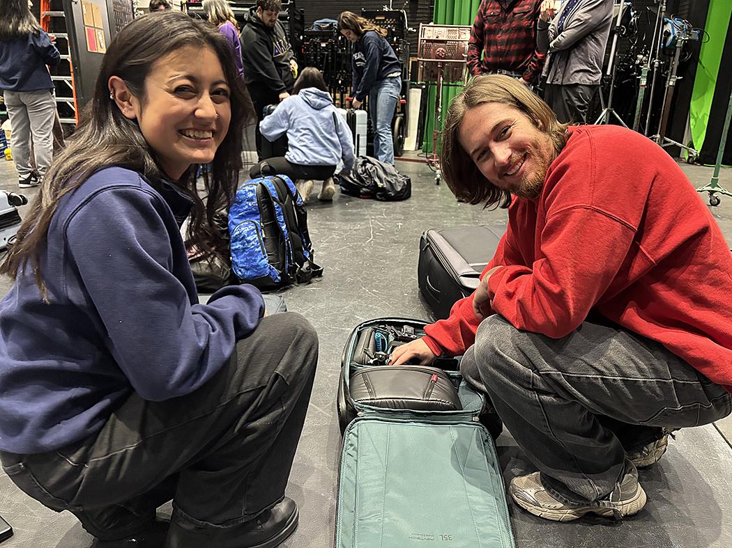 Two kneeling students, a girl with long hair in a blue sweatshirt and a man with a beard in a red sweatshirt
