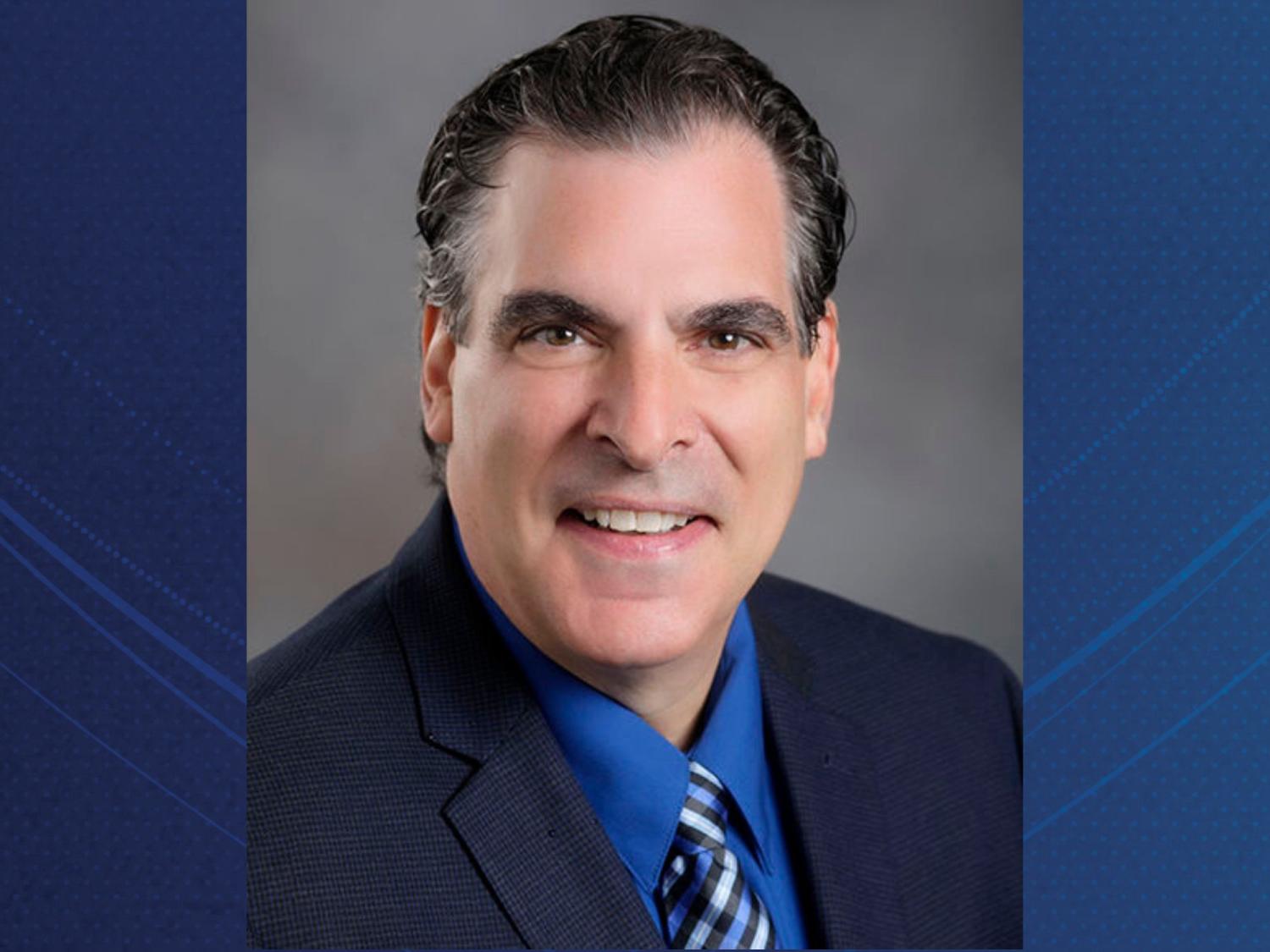 Photograph of a man wearing a dark suit jacket, blue dress shirt, and striped tie. 