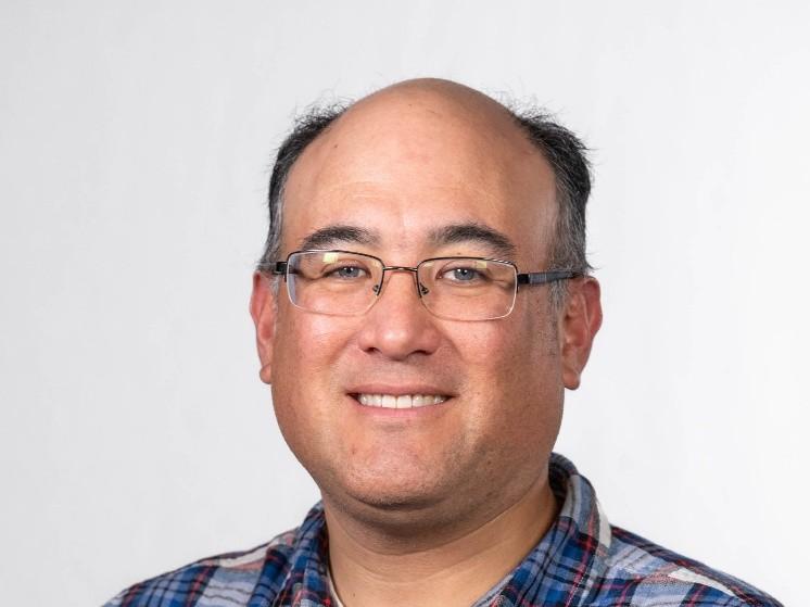 Photograph of a smiling man with glasses wearing a blue, white, and red plaid shirt.