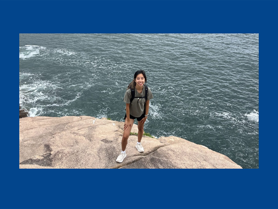 A person, Lydia Neuhauser, is wearing a brown t-shirt and shorts stands on a rock with a backpack and water behind them.