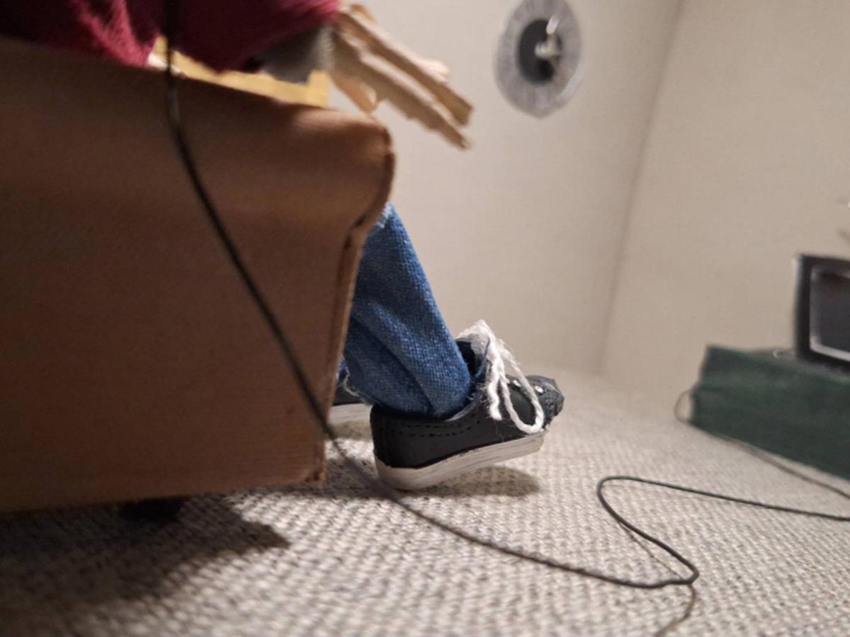 Low-angle view of a small figure sitting on a couch, legs extended, with a TV in the background, creating a playful, miniature-scale scene.