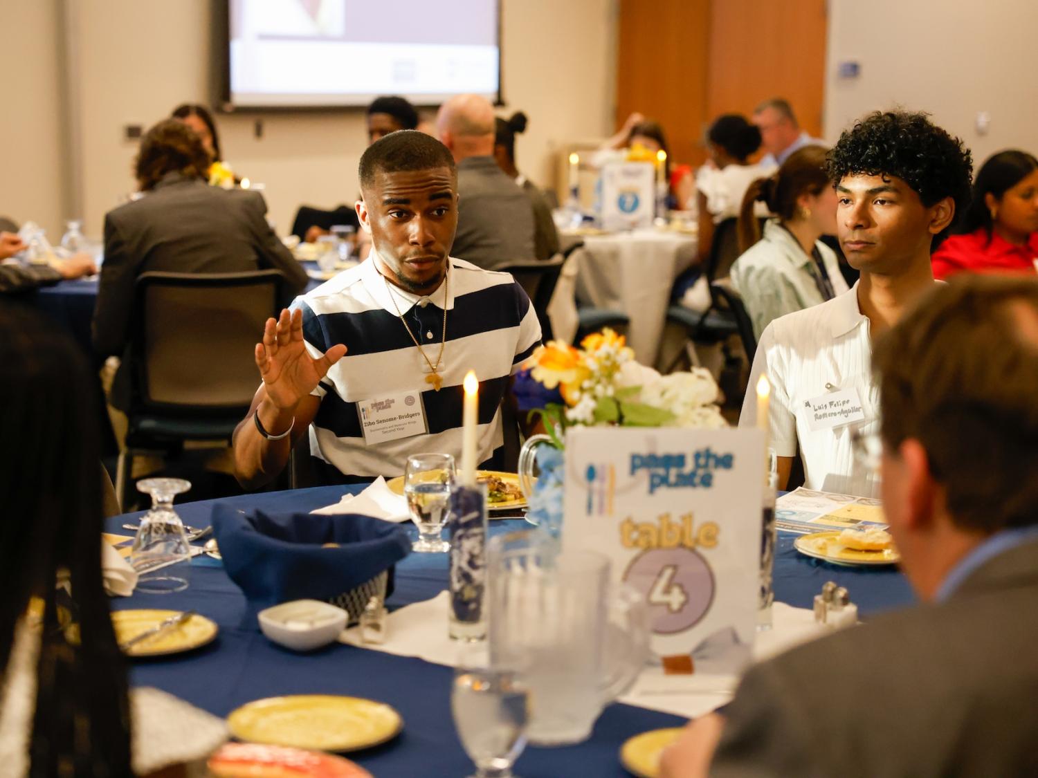 student talking at a table with blue tablecloth and "pass the plate table 4" sign at the table