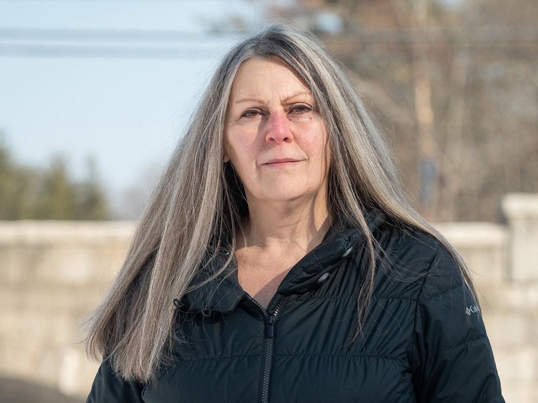 Headshot of Laurene Allen, a woman with long gray hair wearing a black down jacket and standing outside with trees in the background.