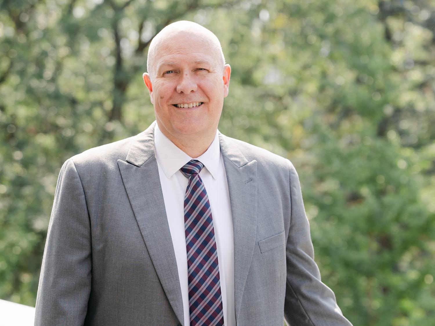 Robert Bohrer wearing a coat and tie outside with trees in the background.