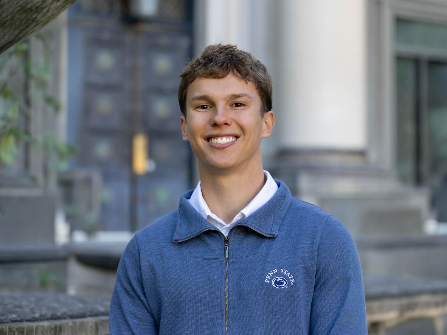 Kyle Sebelin in a light blue quarter zip reading “Penn State” standing outside Sparks building on campus.