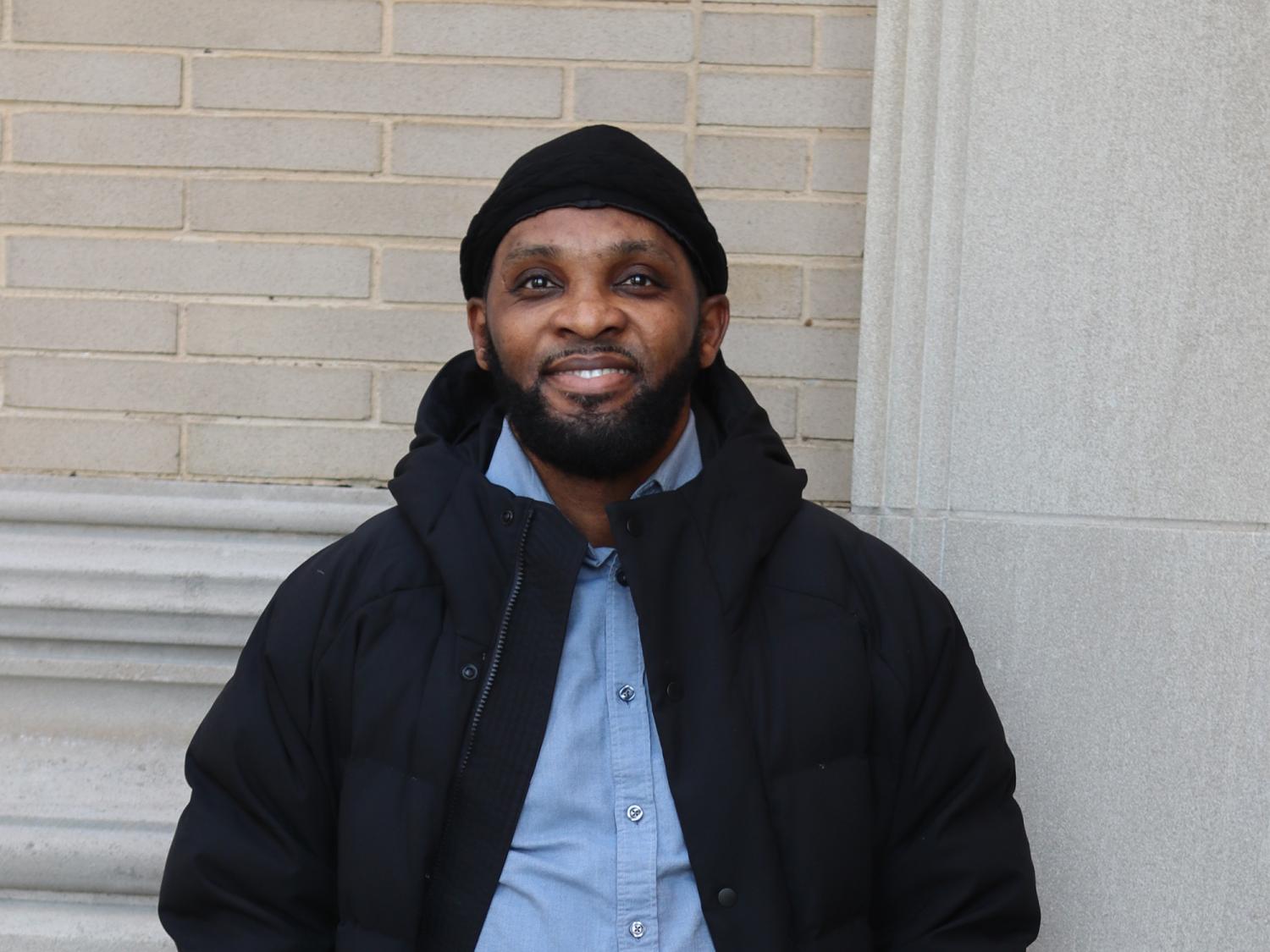 A man wearing a blue shirt and black jacket stands in front of a tan brick wall.