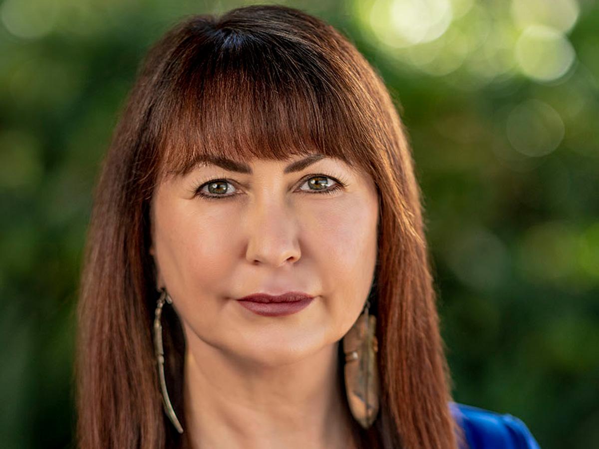 Shannon speed has long brown hair and bangs wears a blue top and large earrings, standing against a softly blurred green background. 