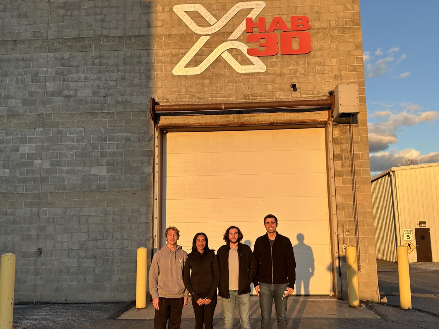A photo of four students standing in front of warehouse garage door with a logo above that reads X-Hab 3D