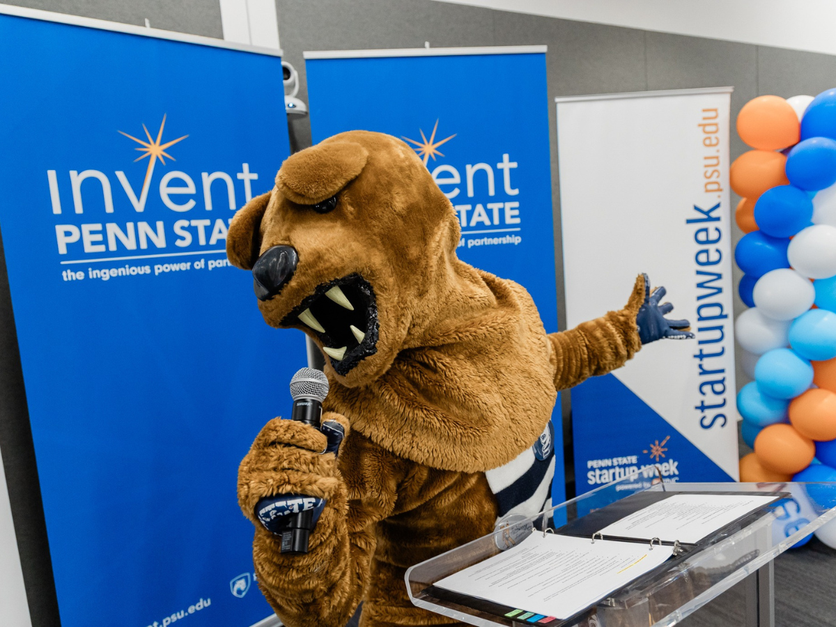 The nittany lion holding a microphone in front of Invent Penn State signage and balloons