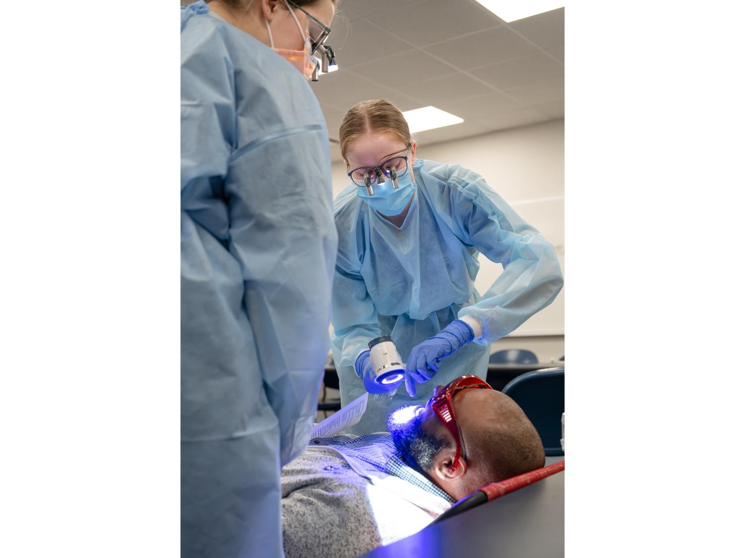 Two students help examine the oral health of an older gentleman on an examination table