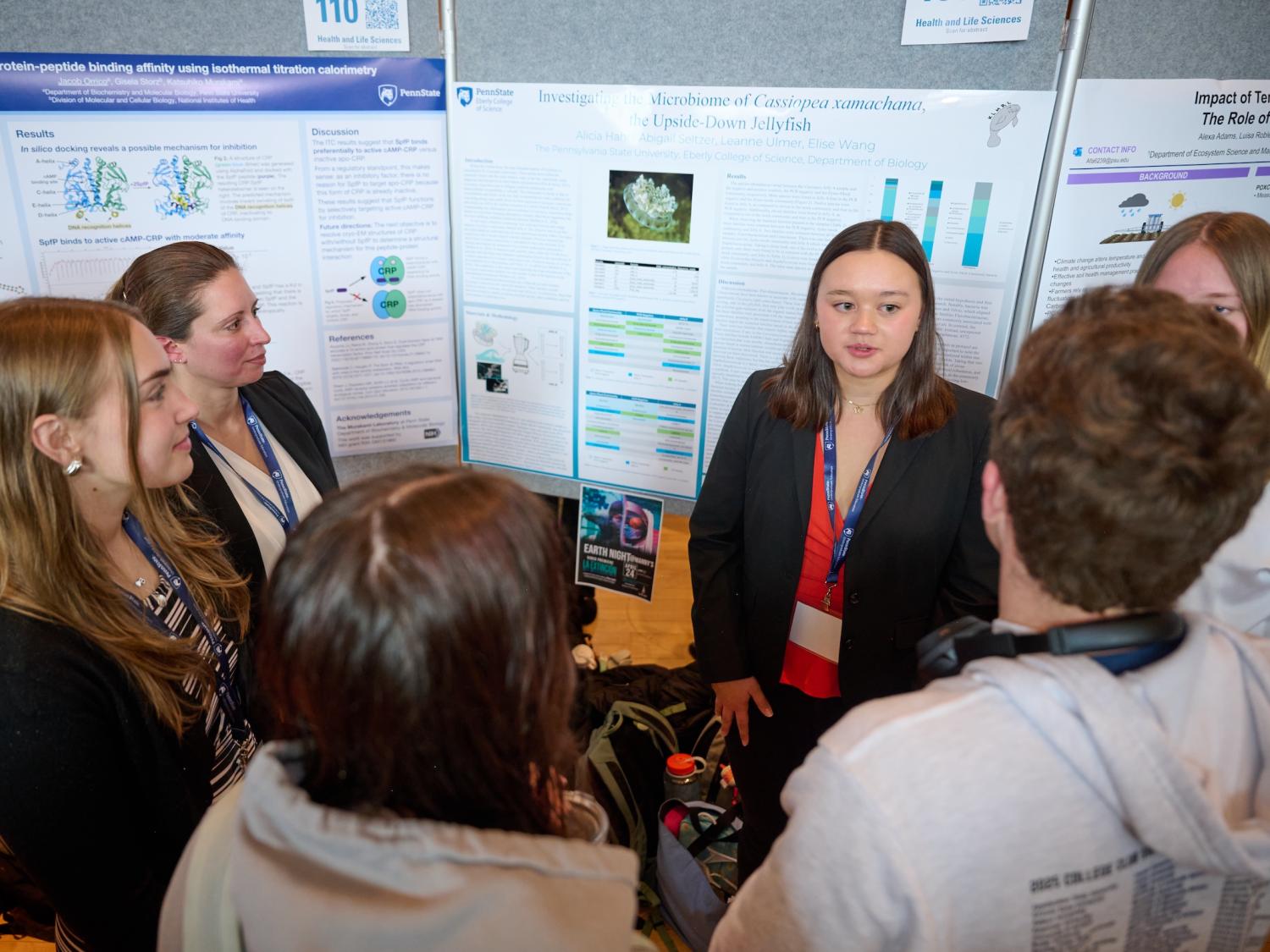 Student presenter talking to half-circle of people in front of research poster