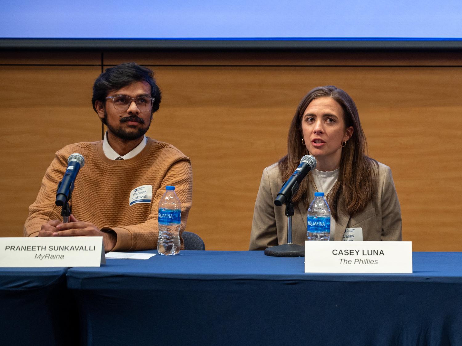 A man and a woman sit behind a table with microphones on it as part of a panel.