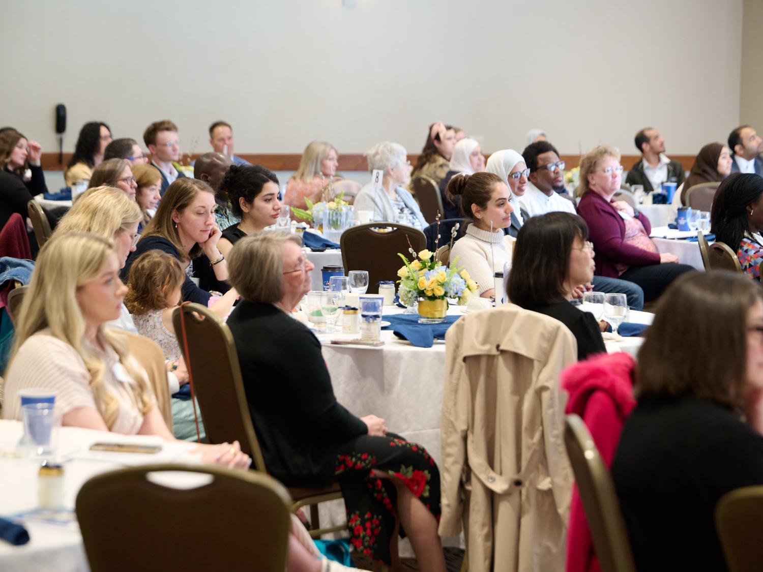 Diverse groups of people sit at round tables with floral centerpieces during a formal indoor event.