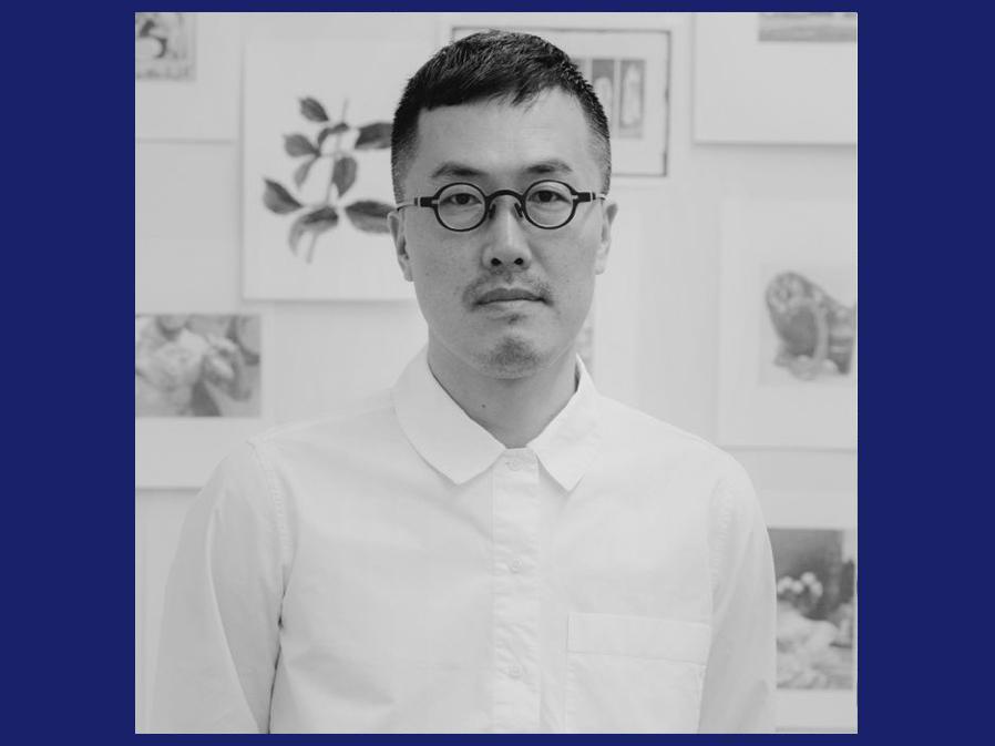 Black and white portrait photo of a person wearing a white shirt with dark hair and dark-rimmed glasses.