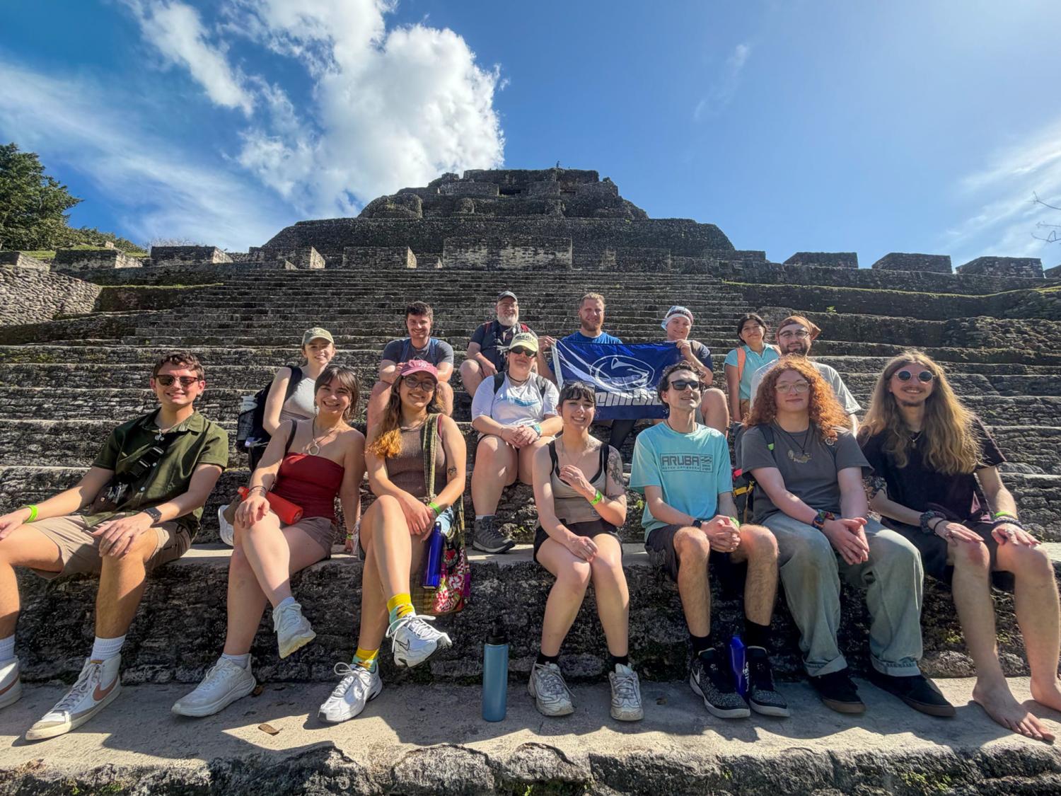 A group of fifteen people sit together on the stone steps of an ancient pyramid at an archaeological site. One person in the back row holds a blue-and-white Penn State flag. The sky is bright and clear, and the group is dressed in casual warm weather clothing.