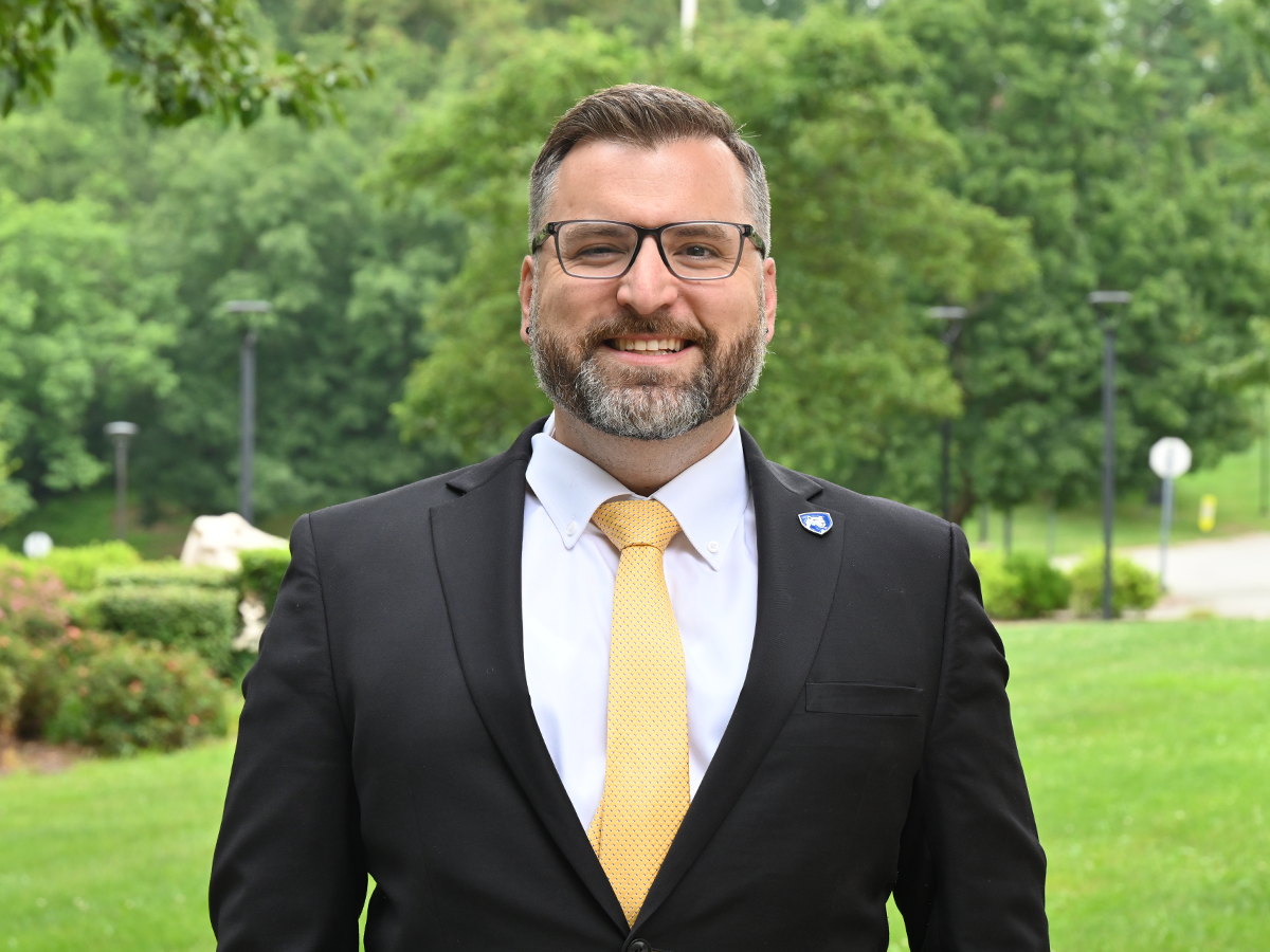 Barrett Scroggs stands outdoors on the Penn State Greater Allegheny campus, smiling at the camera in a black suit, white shirt and gold tie, with trees and campus grounds in the background.