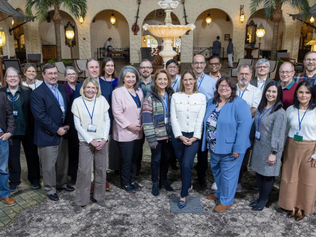 A group of approximately 23 people in business casual attire pose for a group photo in a hotel lobby. A fountain is in the near background.